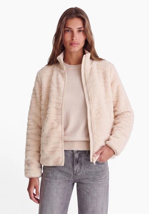 Frau trägt eine cremefarbene, pelzige Jacke über einem beigen Pullover und grauen Jeans, steht mit einer Hand in der Tasche vor einem schlichten weißen Hintergrund.