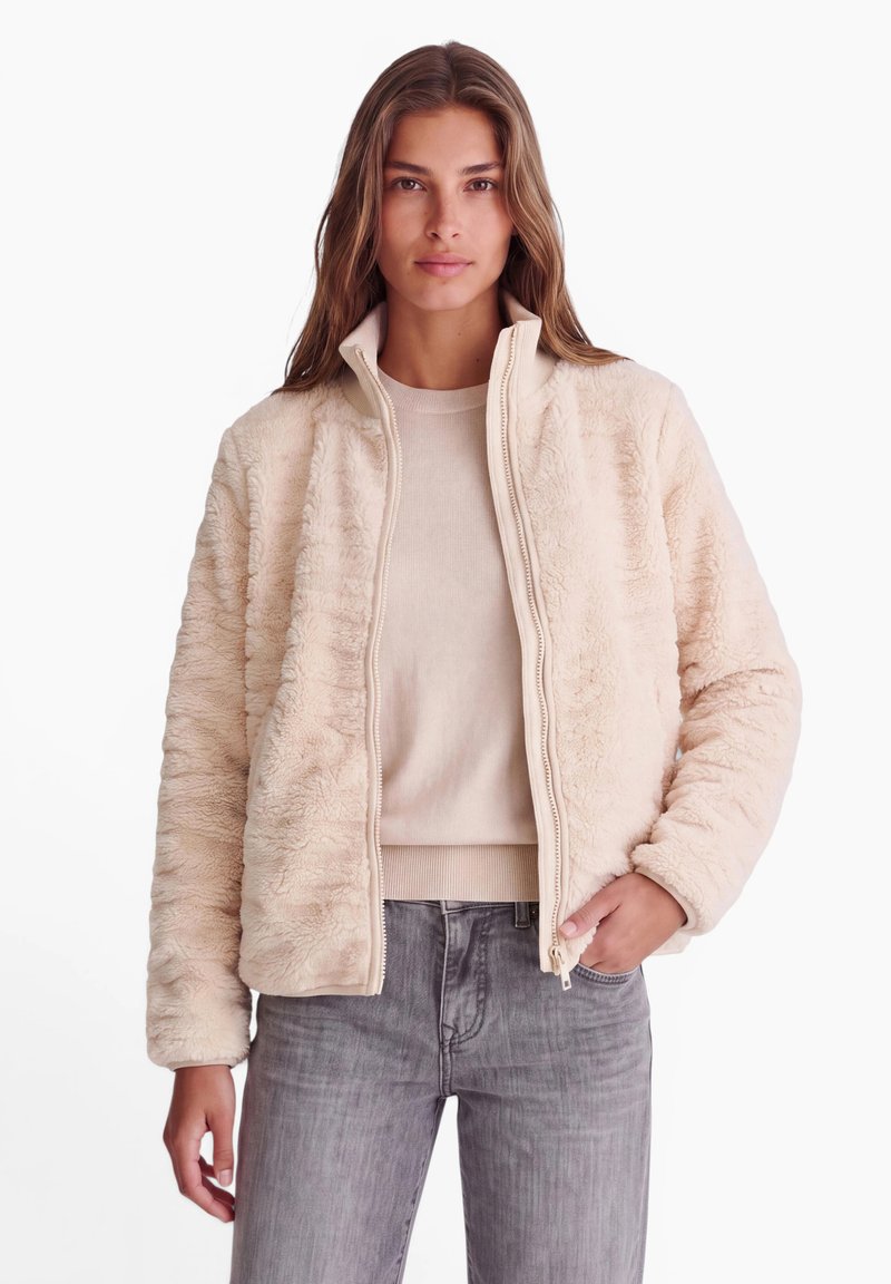 Femme portant une veste duveteuse couleur crème sur un pull beige et un jean gris, debout avec une main dans la poche devant un fond blanc uni.