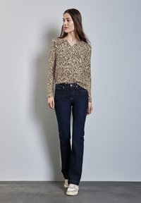 Beige gemusterter Langarm-Top mit einem V-Ausschnitt kombiniert mit dunkelblauen Schlagjeans und beigefarbenen Sneakers auf einem grauen Boden vor einer hellen Wand.