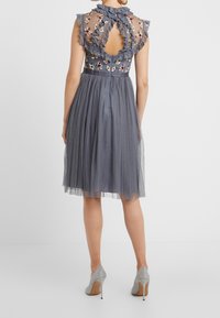 Robe grise avec un corsage transparent brodé de fleurs, détails à volants, dos ouvert et jupe en tulle à plusieurs niveaux, associée à des escarpins argentés.