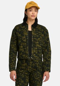 Giacca corta in tessuto floreale verde scuro con colletto nero, dotata di tasche frontali e chiusura con zip. Indossata con un top nero.