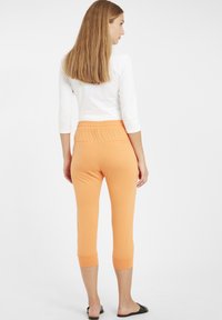 Pantalons capri orange avec une taille élastique et deux poches arrière, associés à un haut à manches longues blanc. Tissu lisse et léger.