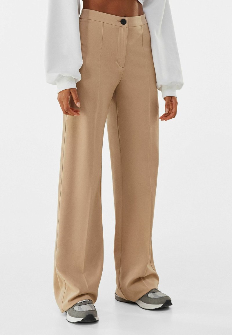 Pantalon beige taille haute à jambes larges avec un bouton noir, porté avec des baskets grises et un top court blanc à manches longues.