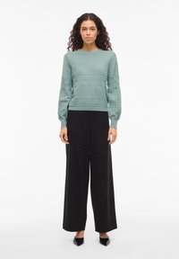 Vrouw staat frontaal en draagt een lichtgroene trui met textuur, zwart wijde pijpen broek en zwarte pumps met puntige neus tegen een witte achtergrond.