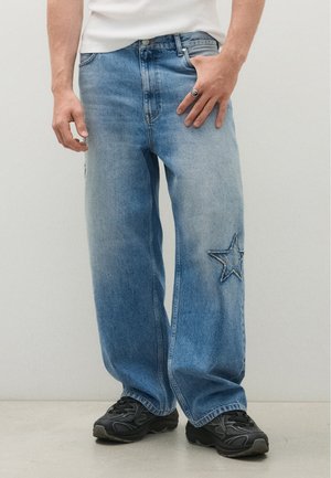 Personne portant un jean bleu clair ample avec une étoile cousue sur une jambe, des baskets noires et une chemise blanche, debout devant un fond uni.