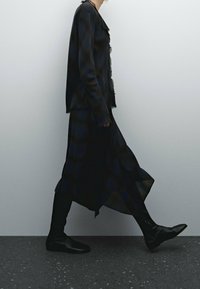 Robe longue à carreaux noirs avec une bordure effilochée, associée à un blazer noir assorti et des bottes noires jusqu'au genou, sur un fond neutre.