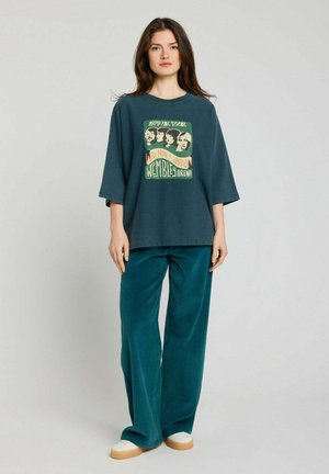 Jeune femme debout portant un t-shirt oversize vert foncé de groupe, un pantalon large bleu sarcelle et des baskets blanches sur un fond uni.