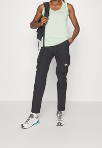 Ljust grönt ärmlöst linne, svarta cargobyxor med sidofickor och flerfärgade träningsskor. Ryggsäck bärs över axeln.