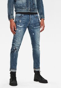 Bleka blå denimjeans med slitna detaljer, färgstänk och smal passform. Utrullad nederkant och ihopparade med svarta snörkängor.