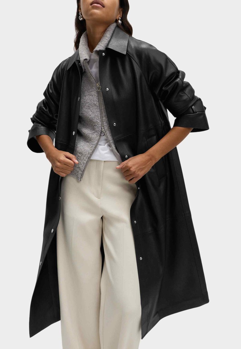 Černý kožený dlouhý kabát volného střihu s knoflíkovým zapínáním, širokým límcem a ohrnutými rukávy přes šedou zipovou bundu a krémové kalhoty.