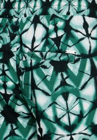 Vihreä kuvioitu kangas, jossa on geometrisia muotoja vaalean ja tumman vihreän sävyjen sekoituksella. Mukana tasku, jossa on metallinen merkitagsi.