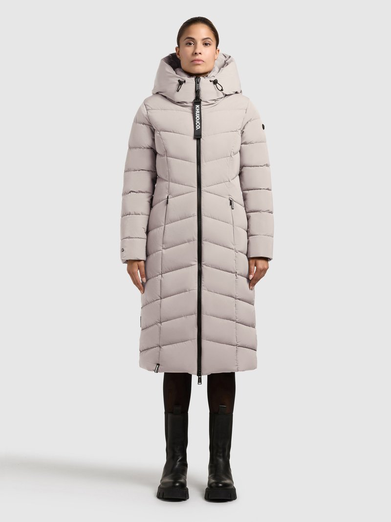 Dlhá svetlosivá prešívaná bunda s kapucňou a čiernym zipsom. Má vzor chevron, bočné vrecká a logo na prednej strane.