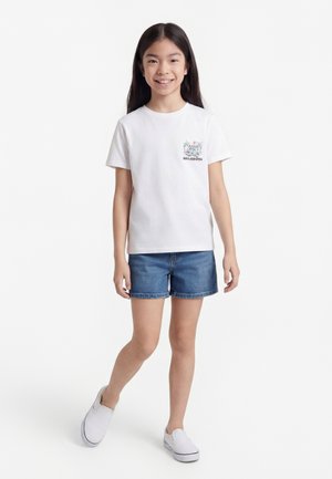 Smilende pige med langt mørkt hår iført hvid grafisk t-shirt, denimshorts og hvide slip-on sko, stående på ét ben.