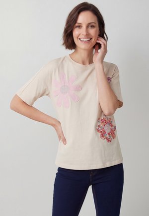 Lächelnde Frau mit kurzen braunen Haaren, die ein beiges T-Shirt mit pinken und floralen Blumenmustern trägt und dunkelblaue Jeans sowie mit einer Hand in der Hüfte posiert.
