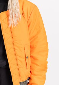 Oranje pufferjack met een doorgestikte textuur, voorzien van een zijzak met een drukknop. De manchetten hebben een contrasterende grijze accent.