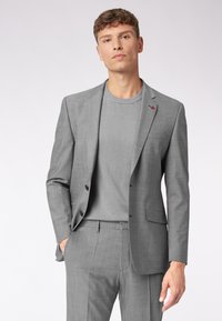 Grau Anzug mit taillierter Passform, strukturiertem Stoff, einreihigem Sakko und einem Rundhals-Grauen Pullover darunter. Dezent roter Akzent.