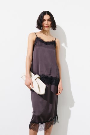 Nainen, jolla on lyhyet tummat hiukset ja joka pitää kädessään taiteltua valkoista clutch-laukkua valkoista seinää vasten, hänellä on yllään hihaton tumma satiinimekko, jossa on mustaa pitsiä.