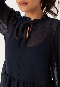 Blusa strutturata di un blu navy, con un collo alto e chiusura a cravatta, motivo a pois e bottoni frontali. Maniche lunghe trasparenti e design a strati.