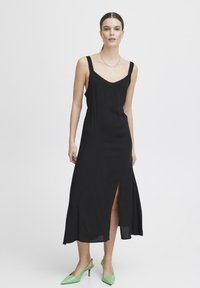 Fekete pántos maxi ruha V-nyakkivágással, oldalhasítékkal, sima textúrával; zöld, hegyesorrú, slingback cipőkkel párosítva.