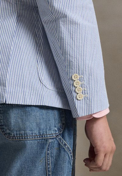 Svetlomodrá a bielo pruhovaná bunda so štyrmi béžovými gombíkmi na manžete. Má vrúbkovaný klopový golier a priliehavý strih. Nosená cez denim.