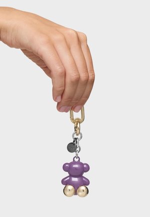 Hand hält einen Schlüsselring mit einem lila, bärenförmigen Anhänger mit goldfarbenen Füßen und einem kleinen runden Etikett.