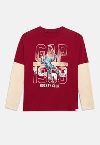 Gyermek rétegezett, vörös és bézs hosszú ujjú ing "GAP 1969 Hockey Club" felirattal és jégkorongozót ábrázoló grafikával, aki bottal tart egy korongot.