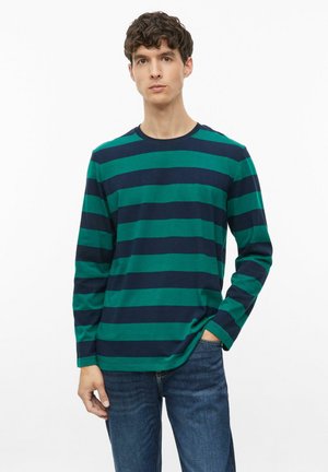Jonge man die een groen en navy blauw horizontaal gestreept shirt met lange mouwen en een blauwe spijkerbroek draagt, staat met één hand in zijn zak.
