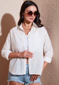 White linen button-up shirt with roll-up sleeves, worn over a white tank top and paired with denim shorts. Accessories include layered gold necklaces and sunglasses.