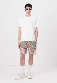 T-shirt blanc à manches courtes, shorts floraux verts avec des feuilles rouges, baskets blanches, chaussettes à hauteur de cheville et lunettes de soleil. Design moderne et simple.