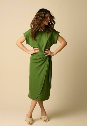 Femme aux cheveux ondulés portant une robe midi verte à manches courtes et des sandales beiges à talons, debout les mains sur les hanches et souriante.