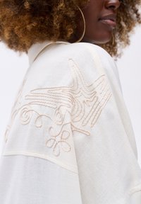 Witte blouse met ingewikkelde beige borduursels met bloem- en abstracte ontwerpen, zachte textuur en een lossere pasvorm.