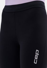 Close-up of black athletic leggings with gray "cep" logo on thigh, worn with a light lavender top.
