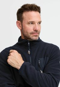 Dunkelblaue Fleecejacke mit hohem Kragen, Reißverschluss vorne und Seitentasche. Sie hat eine weiche Textur und elastische Bündchen.
