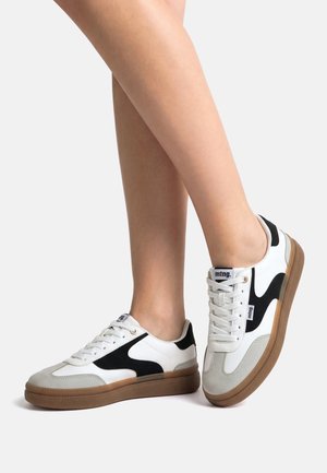 Gambe con scarpe da ginnastica bianche con dettagli neri e suola marrone, un piede appoggiato a terra, l'altro leggermente sollevato sulle punte.