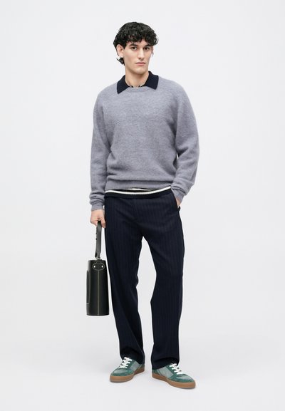 Maglione grigio in lana con colletto blu navy, camicia a righe sotto, pantaloni blu navy a righe sottili e sneakers teal. Tiene una borsa nera.