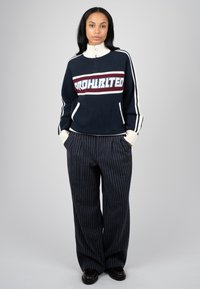 Femme portant un pull bleu marine avec des rayures blanches et bordeaux et le mot "PROHIBITED,", associé à un pantalon large à fines rayures foncé.