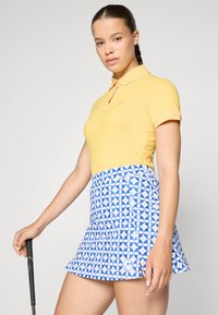 Polo shirt giallo a coste con colletto bianco e gonna blu a fantasia con disegni circolari e un logo sull'orlo.