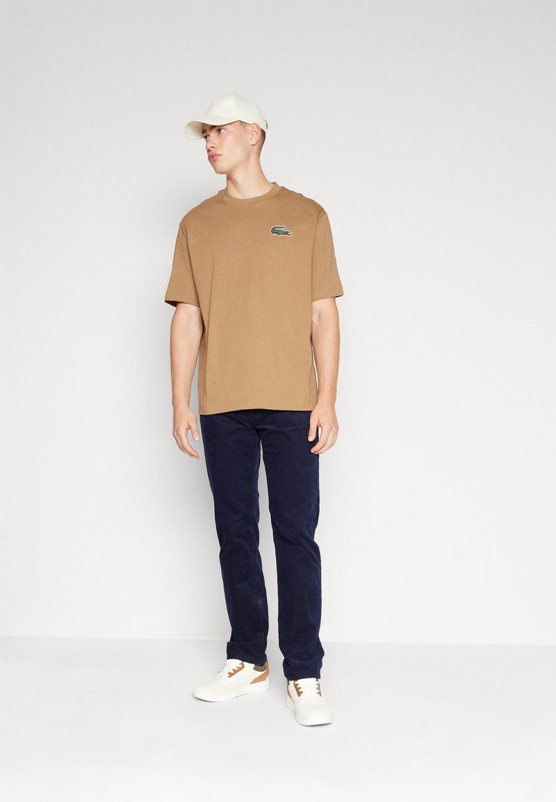 T-shirt di cotone marrone con logo verde sul petto, abbinato a pantaloni blu scuro e sneakers bianche con accenti marroni.