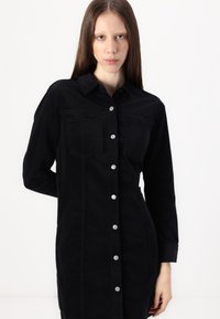 Black denim shirt dress with a button-up front, two chest pockets, long sleeves, and a relaxed fit. Features silver buttons and visible stitching.