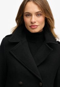 Zwarte double-breasted jas gemaakt van een wolmix, met brede opgestikte revers, gestructureerde stof en metalen knopen. Gestyled over een zwarte coltrui.