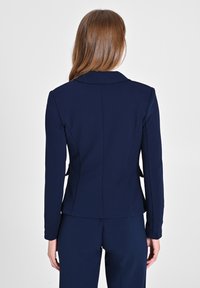 Blazer ajusté bleu marine avec un col, des épaules structurées et une silhouette cintrée. Tissu doux et lisse avec des poches latérales.