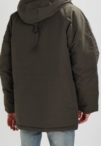 Persona che indossa un giubbotto invernale verde oliva con cappuccio e jeans azzurri chiari, vista da dietro su uno sfondo semplice.