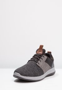 Sneaker sportivo nero con tomaia in rete intrecciata, dettagli grigi, finiture testurizzate, lacci rotondi e suola imbottita con linguetta.