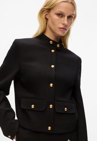 Giacca nera con bottoni dorati, design strutturato, colletto alto, due tasche frontali e texture liscia, che enfatizza una silhouette aderente.