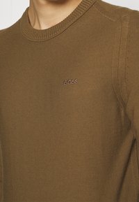 Gros plan sur une personne portant un pull en tricot marron avec un col rond côtelé et le mot "BOSS" brodé sur la poitrine.