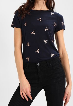 T-shirt blu navy a maniche corte, con un motivo di piccoli uccelli rosa chiaro. Indossato con jeans a righe nere.