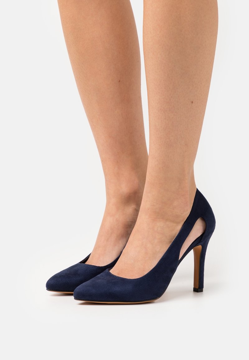 Escarpins en daim bleu marine à talons hauts, avec côtés découpés, bouts pointus et talons aiguilles fins. Texture lisse avec détails de couture ton sur ton.