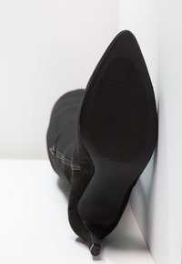 Černé sametové boty na vysokém podpatku se špičatou špičkou a vroubkovanou podešví opřené o bílou zeď a povrch.