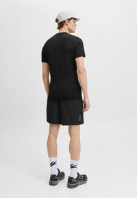 Schwarzes, eng anliegendes T-Shirt kombiniert mit schwarzen Sportshorts. Das Model trägt schwarze Schuhe und gemusterte weiße Socken. Eine hellgraue Kappe rundet das Outfit ab.