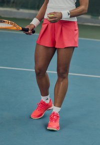 Cortos pantalones de tenis rosas con pliegues, combinados con zapatillas deportivas rosas brillantes y calcetines cortos blancos, sobre una superficie de cancha azul.